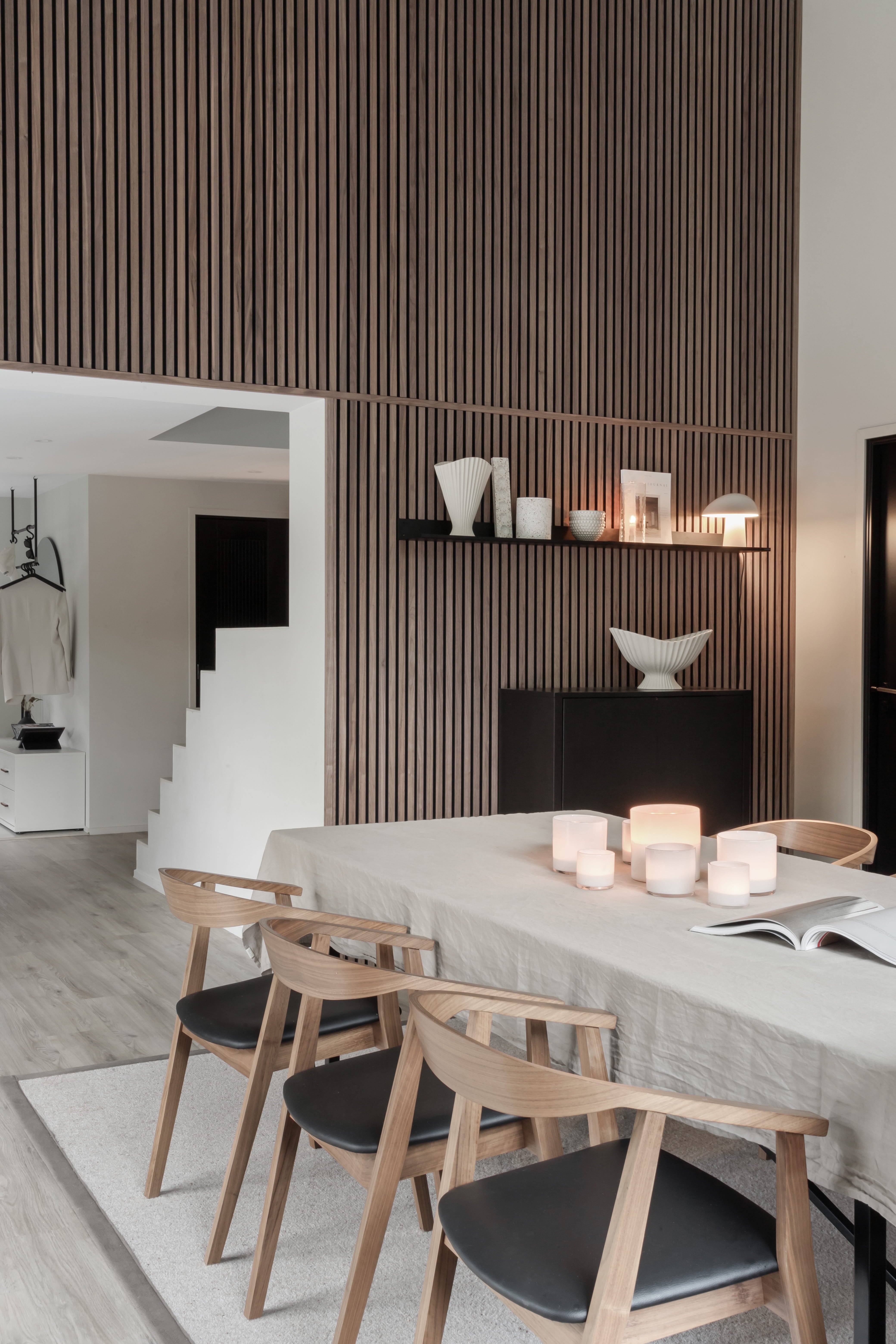 Sala da pranzo moderna con tavolo in legno chiaro, quattro sedie, candele e arredamento minimalista. Parete verticale in doghe acustiche Akupanel e scala a vista.